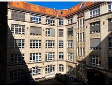 Mehrstöckiges Backsteingebäude mit heller Fassade und zahlreichen weißen Sprossenfenstern, die einen Innenhof umschließen. Einige Fenster haben heruntergelassene Jalousien, und im Hof stehen Mülltonnen sowie ein parkendes Auto.