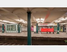 Unterirdischer U-Bahnhof Wittenbergplatz mit grün gestrichenen Stahlstützen, einer kleinen verglasten Kabine im linken Bereich, einem Stationsschild, roten Sitzbänken und Plattformen. Die Beleuchtung erfolgt durch helle Deckenlampen, und Gleise verlaufen im Vordergrund.