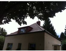 Einfamilienhaus mit hellem Putz und rotbraunem Ziegeldach, mit Dachgaube und Schornstein. Das Gebäude liegt in einem grünen Garten, oben von einer großen Baumkrone überragt und von hohen Nadelbäumen umgeben.