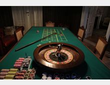Ein Roulette-Tisch in einem klassisch eingerichteten Raum mit grüner Spielfläche, Roulettekessel, Einsatzchips und Rechen. Im Hintergrund stehen gepolsterte Stühle und eine Tür, die zu einem angrenzenden Bereich führt.