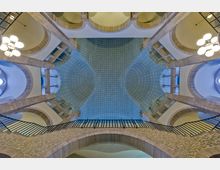 Blick nach oben in einen historischen Innenraum mit symmetrischen Bögen, sandfarbenen Steinsäulen und einem geschwungenen, blauen Deckengewölbe, das mit weißen Mustern verziert ist. Im Vordergrund ist ein Geländer aus Metall zu sehen, das eine Balustrade begrenzt.