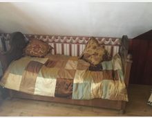 Holzbankbett mit geschnitzten Armlehnen, bedeckt mit einer Patchwork-Decke in warmen Braun-, Gold- und Grüntönen sowie zwei dekorativen Kissen. Die Wand dahinter ist mit gestreifter Tapete und einer Bordüre mit floralem Muster gestaltet.