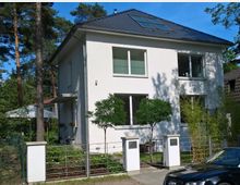 Zweistöckiges, modernes Einfamilienhaus mit weißer Fassade und einem dunklen Schrägdach mit Dachfenstern, umgeben von einem gepflegten Garten mit Bäumen und einem Metallzaun. Im Vordergrund ist ein gepflasterter Gehweg sowie ein Auto zu sehen, während im Hintergrund Bäume und weitere Gebäude stehen.