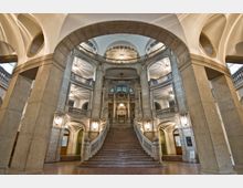 Innenansicht eines prunkvollen Gebäudes mit einer großen, zentralen Steintreppe, die zu einer verzierten Galerie führt. Der Raum ist von hohen Säulen, Rundbögen und dekorativen Balustraden geprägt, mit kunstvollen Deckenleuchten und symmetrischer Architektur.