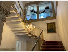 Innenansicht eines historischen Treppenhauses mit weißer, geschwungener Treppe, goldfarbenem Geländer und rotem Teppichläufer. Ein Kristalllüster hängt im Luftraum, darüber sind große Rundbogenfenster und an der Wand ein gerahmtes Gemälde zu sehen.