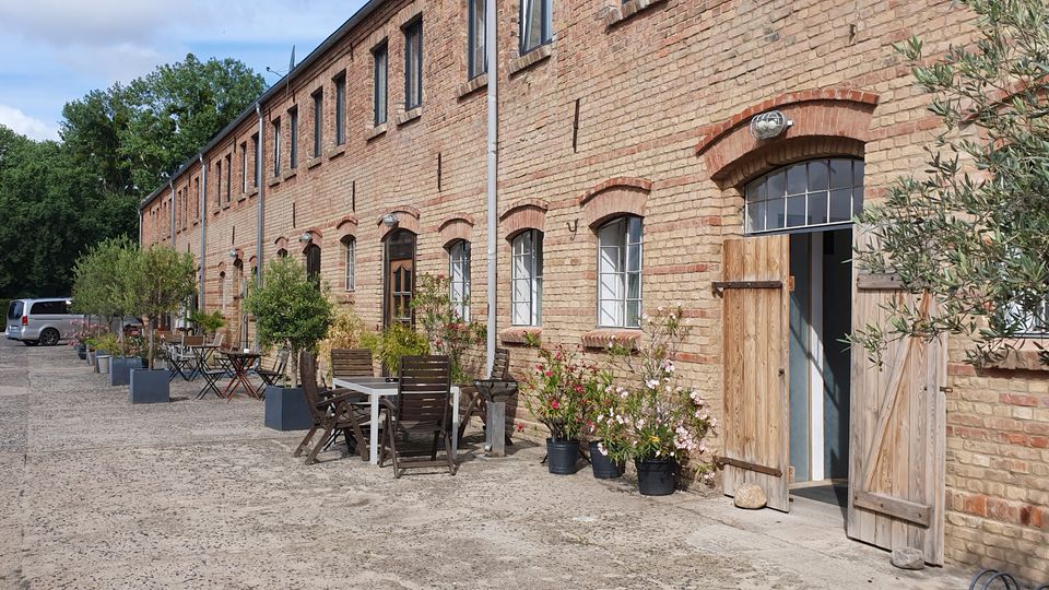 Langer Backsteinbau im Industriehof mit hohen Rundbogenfenstern und geöffneten Holztoren entlang der Fassade. Davor stehen auf der breiten Betonfläche mehrere Tische und Stühle sowie Kübelpflanzen, im Hintergrund sind Bäume und geparkte Fahrzeuge zu sehen.