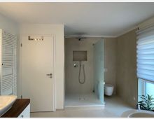 Modernes Badezimmer mit einer bodengleichen Dusche, einer halbtransparenten Glasabtrennung, einem weißen Wand-WC und einem großen Fenster mit Rollos rechts. Links sind ein Heizkörper, eine Tür und ein Waschtisch mit Holzelementen sichtbar.
