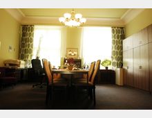 Ein Büro oder Besprechungsraum mit einem Holztisch und umstehenden gepolsterten Stühlen in der Mitte, zwei große Fenster mit Vorhängen im Hintergrund, sowie Schränke und Pflanzendekoration im Raum. Eine Deckenleuchte mit mehreren Lampenschirmen hängt zentral über dem Tisch.