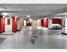 Ein heller, sauberer Tiefgaragenbereich mit rot gestrichenen Wänden und weißer Decke. Es sind Parkplätze, markierte Pfeile auf dem Boden, ein geparktes weißes Auto sowie Türen und Hinweise zu einem Ausgang in der Ferne sichtbar.