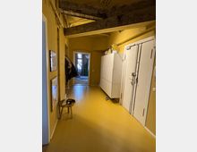 Long, narrow interior hallway with yellow walls and floor, exposed wooden beams overhead, and a doorway at the far end leading to a brighter room. White storage cabinets and a large white door line the right wall, with framed pictures and a small stool along the left side.