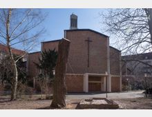 Außenansicht einer modernen Backsteinkirche mit schlichtem Design, einem großen Kreuz an der Hauptfassade und einem kleinen Turm mit Glockenfenstern. Im Vordergrund sind Bäume, Laternen und ein kleiner Sandkasten in einem parkähnlichen Umfeld sichtbar.