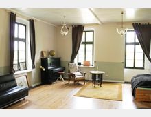 Ein Wohnraum mit Holzboden und mehreren Sprossenfenstern, die von langen dunklen Vorhängen eingerahmt sind. Im Raum befinden sich ein schwarzes Klavier, ein Sofa, ein Sessel, ein kleiner runder Tisch und ein Einzelbett; die Einrichtung wirkt klassisch und gemütlich.