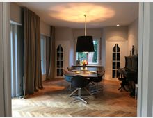 Esszimmer mit modernen Möbeln, bestehend aus einem Esstisch mit mehreren Stühlen und einer großen schwarzen Hängelampe darüber. Im Hintergrund befinden sich ein Fenster, zwei Vitrinenschränke mit Glastüren und ein schwarzes Klavier; der Raum hat Parkettboden und helle, bodenlange Vorhänge.