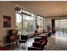 Ein heller Raum mit großen Fenstern, die Tageslicht hereinlassen, ausgestattet mit farbigen Retro-Sesseln, kleinen Tischen und einem alten Radio. Im Hintergrund sind ein Filmprojektor sowie Plakate und eine Aussicht auf Bäume sichtbar.