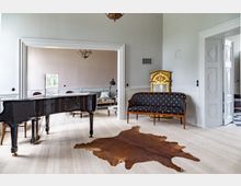 Klassischer Innenraum mit einem schwarzen Flügel, einer goldenen Standuhr, einer gepolsterten Sitzbank mit Mustern und einem braunen Fellteppich auf hellem Holzboden. Im Hintergrund befindet sich ein angrenzender Raum mit einem Sofa und einem kleinen Fenster.