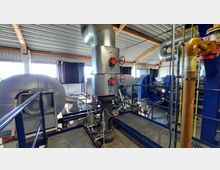 Innenansicht einer technischen Anlage mit Metallrohren, Lüftungselementen und Maschinen. Im Vordergrund befinden sich silberne und orangefarbene Rohre mit roten Ventilen, im Hintergrund sind blaue Maschinen sowie eine Dachkonstruktion aus Holz und Metall sichtbar.