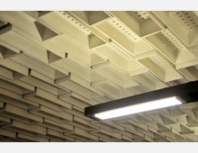 Geometrische Decke mit dreieckigen Vertiefungen und kleinen runden Löchern in einem modernen Innenraum, ergänzt durch eine rechteckige Deckenleuchte in schwarzem Rahmen.