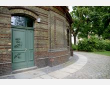 Eingangsbereich eines Backsteingebäudes mit grüner Doppeltür und darüberliegendem Rundbogenfenster, neben einem gepflasterten Weg, der von grünen Bäumen und Büschen gesäumt ist.