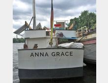 Ein weißes Boot namens "Anna Grace" am Ufer eines Flusses, mit einer am Heck befestigten deutschen Flagge. Im Hintergrund sind Bäume, ein weiteres Boot und ein bewölkter Himmel zu sehen.