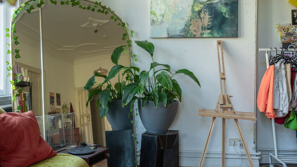Cozy apartment studio corner with a large floor mirror draped in ivy, potted leafy plants on dark stands, and an abstract painting on a white wall. A wooden easel stands beside a small clothing rack, with a sofa edge and record player visible in the foreground.