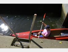 Ein Mann sitzt in einem roten Kajak mit Paddel auf einem dunklen Wasserkanal, umgeben von einem Betonufer mit metallischem Geländer. Im Wasser treiben vereinzelt Blätter, und Sonnenlicht spiegelt sich auf der Oberfläche.