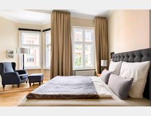 Helles Schlafzimmer mit einem großen Bett mit gepolstertem Kopfteil, einer grauen Decke und Kissen. Der Raum hat große Fenster mit beigefarbenen Vorhängen, einen blauen Sessel mit passendem Hocker sowie helle Wände und Holzfußboden.