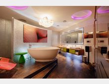 Modernes Badezimmer mit einer freistehenden ovalen Badewanne auf einem erhöhten Podest, einer großen Spiegelwand mit integrierter Beleuchtung und zwei Waschbecken. Die Wandgestaltung enthält glänzende Mosaikfliesen, ein abstraktes Kunstbild und Deckenelemente mit violetter Akzentbeleuchtung.