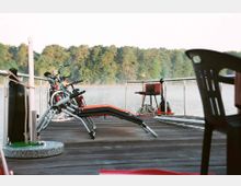 Holzsteg mit Liegestühlen, Fahrrädern und einem Tischgrill, der auf einem ruhig gelegenen Gewässer endet. Im Hintergrund sind nebelverhangene Bäume am Ufer zu sehen.