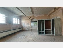 Ein leerer, heruntergekommener Raum mit abblätternder Wandfarbe, zwei Fenstern mit vergitterten Scheiben auf der linken Seite, offenen und beschädigten Schränken rechts, sowie einer Türöffnung in der Mitte, die in einen weiteren Raum führt. Der Boden ist staubig, und die Decke zeigt alte Rohrleitungen.