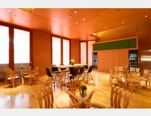 Moderner Gemeinschaftsraum mit Holzwänden und -böden, ausgestattet mit runden Glastischen, Holzstühlen und einer größeren Tischgruppe mit schwarzen Kunststoffstühlen. Im Hintergrund befinden sich bodentiefe Fenster mit milchigen Scheiben, Getränkekühlschränke und eine grüne Wanddekoration.