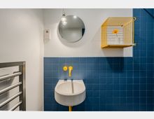 Modernes Badezimmer mit wandhängendem weißem Waschbecken vor blauen Fliesen und gelben Armaturen. Darüber hängen ein runder Spiegel mit Lampe sowie ein gelbes Wandregal; links ist ein Handtuchheizkörper zu sehen.