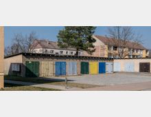 Reihe von Garagen mit bunt gestrichenen Türen in verschiedenen Farben wie Grün, Blau und Gelb vor einem gepflasterten Hof; im Hintergrund befinden sich mehrstöckige Wohnhäuser mit roten Ziegeldächern und kahlen Bäumen.