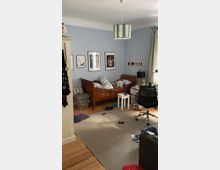 Ein unaufgeräumtes Kinderzimmer mit hellblauen Wänden, an denen gerahmte Bilder und Flaggen hängen. Im Raum befinden sich ein Holzbett mit Kissen, ein Schreibtischstuhl, ein kleiner weißer Tisch sowie Spielzeug und Kleidung auf einem beigen Teppichboden.