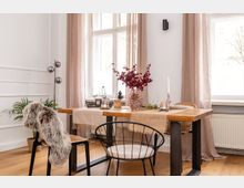 Essbereich mit einem Holztisch und modernen Metallstühlen, dekoriert mit einer Glasvase voller roter Trockenblumen und beige Tischläufern. Helle Vorhänge rahmen große Fenster, und eine Stehleuchte sowie eine Topfpflanze ergänzen die Einrichtung auf einem Holzboden.