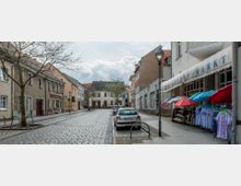 Eine gepflasterte Straße in einer Kleinstadt mit Wohnhäusern und Geschäften. Auf der rechten Seite befindet sich ein Geschäft mit einer Markise, unter der bunte Sonnenschirme und Kleiderständer stehen, während links parkende Autos und ein Fahrradständer zu sehen sind.