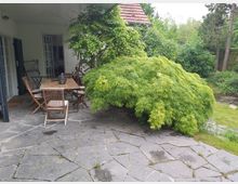 Terrasse mit grauem Steinboden, darauf ein Holztisch mit mehreren Stühlen. Links befindet sich ein überdachter Bereich mit weißen Fenster- und Türrahmen, rechts ein dichter grüner Strauch, umgeben von Bäumen und Rasen im Hintergrund.