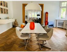 Helles Esszimmer mit einem weißen Tisch, umgeben von grauen und orangefarbenen Stühlen auf einem Holzboden mit Fischgrätenmuster. Im Hintergrund ein goldumrahmter Durchgang, ein rotes dekoratives Element, eine Buddha-Statue und mehrere Fenster mit Blick ins Grüne.