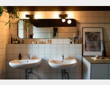 Modernes Badezimmer mit zwei weißen Waschbecken vor einer Wand aus weißen Fliesen. Ein großer, rahmenloser Spiegel, darüber zwei Wandlampen, reflektiert die Umgebung; dekorative Pflanzen und Pflegeprodukte stehen auf dem Fliesensims, und ein gerahmtes Poster steht rechts auf einem Schrank.