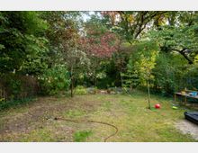 Ein großer Garten mit unebenem Rasen und Erdflächen, umgeben von dichtem Baumbestand und Sträuchern. Im Vordergrund liegt ein Gartenschlauch, rechts stehen ein Tisch mit Gegenständen und ein roter Ball auf der Wiese.