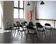 Ein heller Seminarraum mit einer Reihe schwarzer Klappstühle, einer großen Projektionsfläche und einer Boxbirne mit roten Boxhandschuhen. Im Hintergrund sind ein großes Fenster mit Tageslicht sowie minimalistische Hängelampen zu sehen.