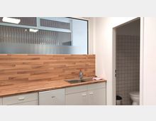 Kleine Küchenzeile mit einer Holzarbeitsplatte, Spülbecken und weißen Schränken, daneben eine Tür zu einem gefliesten Badezimmer mit einer Toilette. Im Hintergrund ist ein Fenster mit Milchglas und einem wellenförmigen Muster zu sehen.