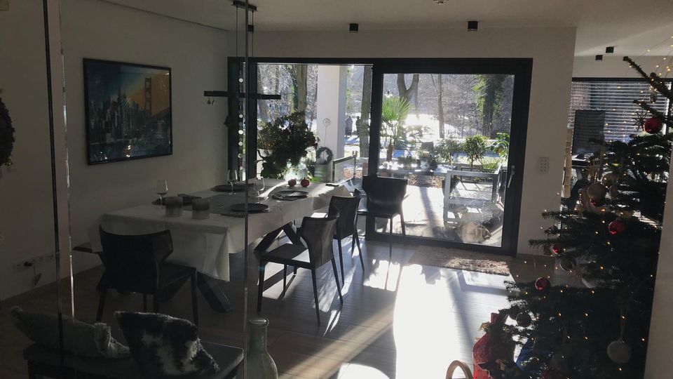 Moderner Essbereich in einem Wohnhaus mit großem Esstisch und dunklen Stühlen, daneben bodentiefen Schiebetüren zur sonnigen Terrasse mit Pflanzen. Rechts steht ein dekorierter Weihnachtsbaum; große Fensterflächen lassen viel Licht in den Raum.