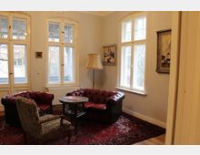 Wohnzimmer mit klassischen Möbeln, darunter zwei lederne Chesterfield-Sessel, ein kleiner Holztisch und ein gepolsterter Sessel mit floralen Mustern. Der Raum hat drei große Fenster mit Sprossen, helle Wände und ist mit einem roten Teppich sowie Gemälden an den Wänden dekoriert.