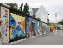 Außenaufnahme eines Abschnitts der Berliner Mauer mit bunten Wandmalereien und Graffiti, die sich entlang einer Straße erstrecken. Im Hintergrund sind moderne Gebäude und der Berliner Fernsehturm zu sehen.