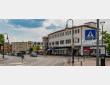 Straßenszene mit einem weißen, zweistöckigen Gebäude mit der Aufschrift „Comédie Soleil“, in dem Geschäfte und ein Drogeriemarkt im Erdgeschoss untergebracht sind. Im Vordergrund ist ein Zebrastreifen und eine leicht kurvige Straße mit mehreren parkenden und fahrenden Autos zu sehen, umgeben von Straßenlaternen und kleineren Gebäuden im Hintergrund.