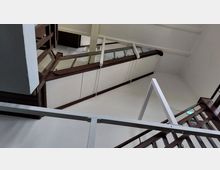 Blick nach oben in einem Treppenhaus mit weißen Wänden und Holzgeländern. Die architektonischen Elemente umfassen mehrere Etagen mit Metallgeländern und diagonalen Trägern.