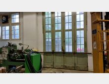 Ein Werkstattraum mit großen, grün lackierten Doppeltüren und Glasfenstern, die von Holzrahmen umgeben sind. Im Vordergrund steht eine grüne Drehmaschine, daneben Kabel und Maschinenteile; rechts ist ein Teil einer gelben Hebevorrichtung sichtbar.