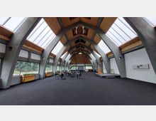 Großer, moderner Innenraum mit einer gewölbten Decke aus Holz und Betonträgern, seitlich große Fenster mit Blick ins Grüne. Der Raum ist mit vereinzelten Tischen, Stühlen und einem kleinen Podium ausgestattet, möglicherweise genutzt für Veranstaltungen.
