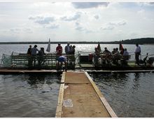 Eine schwimmende Plattform auf einem See, auf der mehrere Menschen stehen oder sitzen. Im Vordergrund führt ein Steg zur Plattform, die mit Tischen, Stühlen und Fahnen ausgestattet ist; im Hintergrund ist die Wasserfläche sowie der von Bäumen gesäumte Horizont sichtbar.
