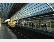 Moderner Bahnhof mit Glasdach; ein Nahverkehrszug steht an einem der Bahnsteige, während Menschen entlang der Bahnsteige gehen oder warten. Auf der rechten Seite steht eine verglaste Wartestation.